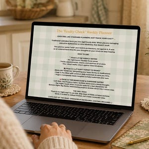 May include: A laptop screen showing the text "The 'Reality Check' Weekly Planner". The laptop is on a wooden table with a cup of coffee, a coaster, a notebook, a pen, and a vase of flowers. The text is in a green and white checkered pattern.