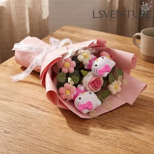 Puede incluir: Un ramo de flores hechas a mano envuelto en papel rosa y atado con una cinta blanca. El ramo incluye flores de ganchillo rosas y blancas y dos figuras de Hello Kitty. La cinta dice "Just For You".