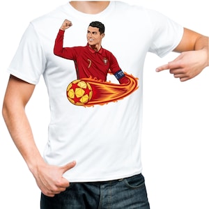 May include: White t-shirt featuring a cartoon illustration of a soccer player in a red jersey with a soccer ball engulfed in flames. The player is celebrating with a raised fist. The design is centered on the shirt.