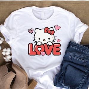 May include: White t-shirt featuring a graphic of a cartoon cat with a red bow, surrounded by hearts and a diamond. The word "LOVE" is displayed in red, with a pink heart in the center of the "O".