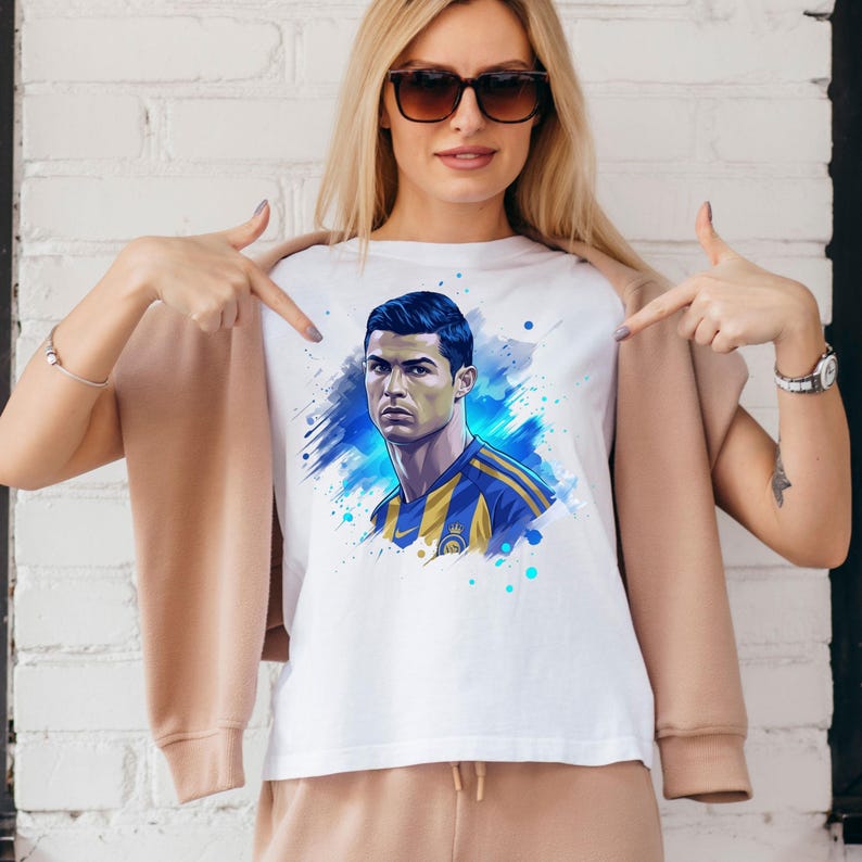 May include: White t-shirt featuring a portrait of a soccer player in a blue and yellow jersey. The graphic has a watercolor effect with blue and teal accents. The shirt is part of a sports apparel collection.