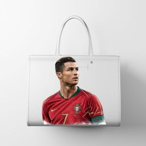 May include: White tote bag featuring a portrait of a soccer player in a red jersey with the number 7. The bag has a leather-like texture and a simple design, with a handle for carrying.