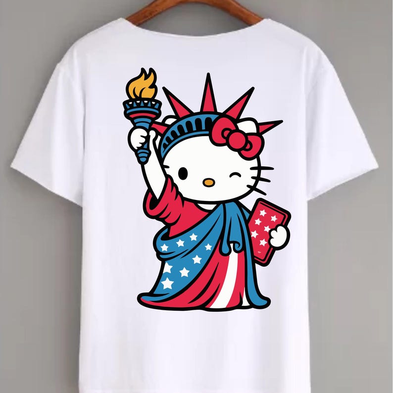 May include: White t-shirt featuring a cartoon illustration of Hello Kitty dressed as the Statue of Liberty. The design includes a red bow, a torch with a flame, and a tablet with stars, all in patriotic colors.