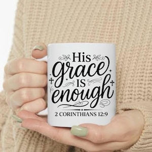 May include: White ceramic mug with the text "His grace is enough" in black script. The mug also features the Bible verse "2 Corinthians 12:9". The mug is held in a person's hands.
