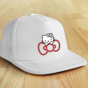 May include: White trucker hat featuring a Hello Kitty graphic. The graphic shows Hello Kitty's face above a red bow. The hat has a white front panel and a white mesh back panel. The brim is curved and white.