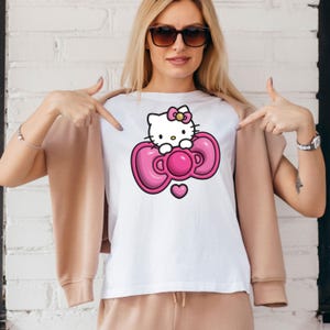 May include: White t-shirt featuring a cartoon image of Hello Kitty. The graphic shows Hello Kitty with a pink bow and a pink bow-shaped design. A small pink heart is below the bow. The shirt is worn by a person.