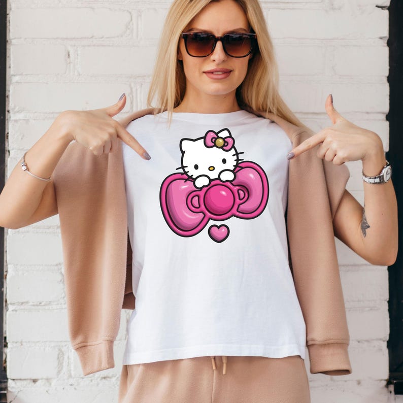 May include: White t-shirt featuring a cartoon image of Hello Kitty. The graphic shows Hello Kitty with a pink bow and a pink bow-shaped design. A small pink heart is below the bow. The shirt is worn by a person.