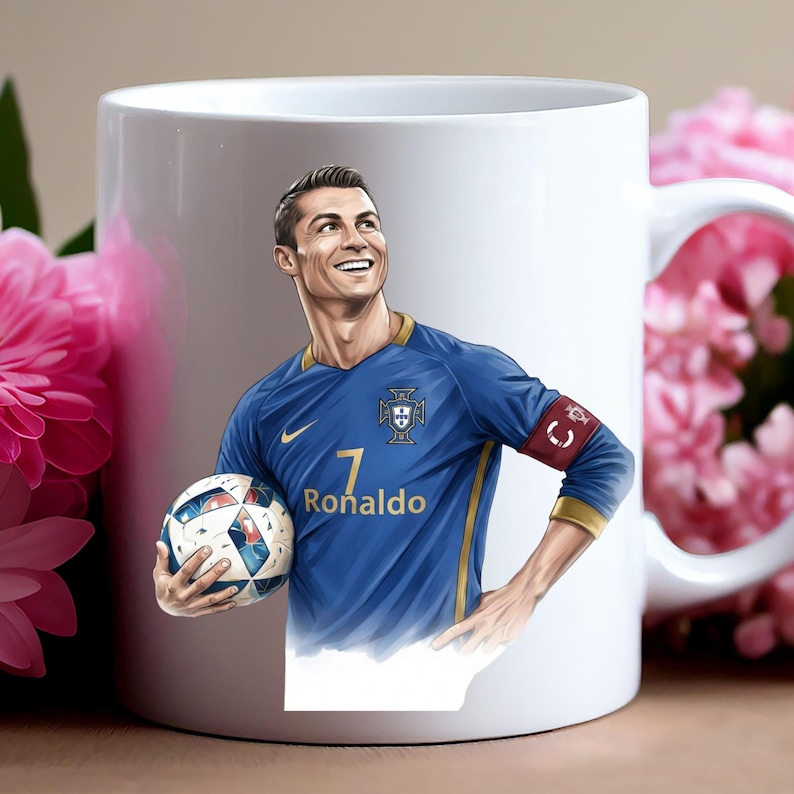 May include: White ceramic mug featuring a portrait of a soccer player in a blue jersey with the number 7 and the name "Ronaldo". The player is holding a soccer ball and smiling. The mug has a handle on the right side.