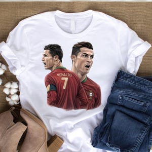 May include: White t-shirt featuring a portrait of Cristiano Ronaldo in a red soccer jersey with the number 7. The shirt is paired with blue jeans and tan boots.