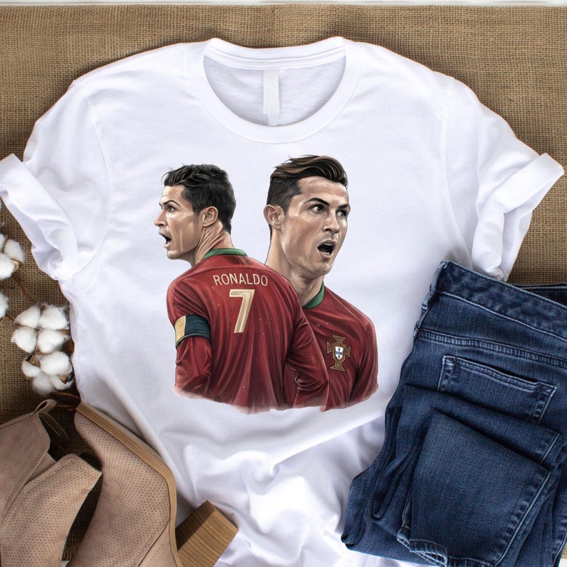 May include: White t-shirt featuring a portrait of Cristiano Ronaldo in a red soccer jersey with the number 7. The shirt is paired with blue jeans and tan boots.