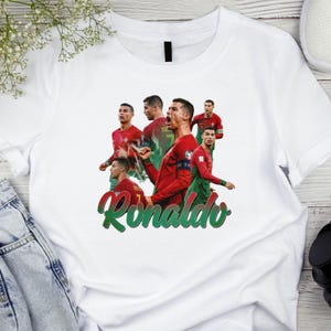 May include: White t-shirt featuring multiple images of a soccer player in a red and green uniform. The word "Ronaldo" is written in green script across the bottom of the shirt.