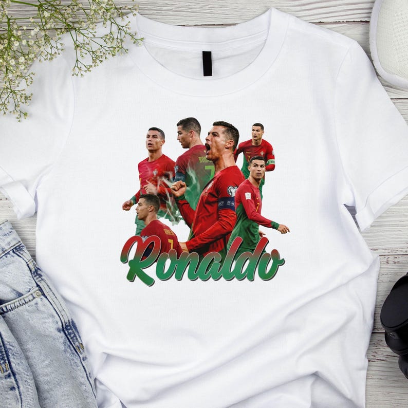 May include: White t-shirt featuring multiple images of a soccer player in a red and green uniform. The word "Ronaldo" is written in green script across the bottom of the shirt.