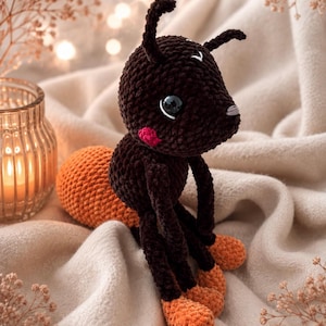 May include: A crocheted ant plush toy featuring a dark brown head and body with a textured surface. The ant has two antennae, large black eyes, small pink cheeks, and an orange abdomen. The legs end in orange feet, matching the abdomen color.