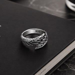 May include: A silver ring with a wing design, marked with "925". The ring is set on a black book with the text "ARTS THE CRAFTS". The ring's wing detail is visible.