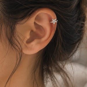 Pode incluir: Um ear cuff de tom prateado com um design em forma de flor adornado com pequenas pedras transparentes de corte marquise. A peça de joalharia está posicionada na cartilagem superior da orelha, exibindo uma banda metálica fina que prende o ornamento floral.