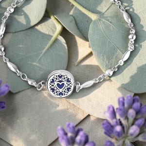 May include: A silver bracelet with a central round charm featuring a blue heart design surrounded by heart-shaped cutouts. The bracelet has a delicate chain with oval and round links. The background includes lavender and eucalyptus leaves.