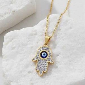 May include: A gold-colored necklace featuring a Hamsa hand pendant. The pendant is encrusted with clear crystals and a blue eye in the center. The necklace is displayed on a white, textured surface.