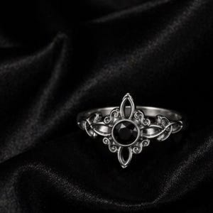 May include: A silver-toned ring featuring a central round black gemstone. The metal band displays intricate, symmetrical scrollwork and filigree patterns that extend outward from the stone, creating a pointed, star-like shape on the front.
