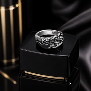 May include: A silver-colored ring with a wing design. The ring is marked with "925" and features detailed feather-like patterns. The ring is displayed on a black surface, with a blurred background.