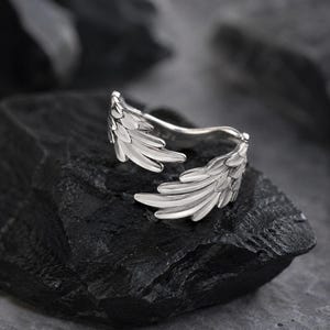 May include: A silver-toned open ring featuring two detailed, overlapping wing designs. The metal surface is polished and reflective, showcasing individual feather textures carved into the band. The ring is shaped to wrap around a finger with an open gap.