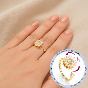 May include: A gold ring with a sunflower design, featuring a central cluster of clear stones and surrounding petals. The ring has a rotating feature and the words "you are my sunshine" are engraved on the band.