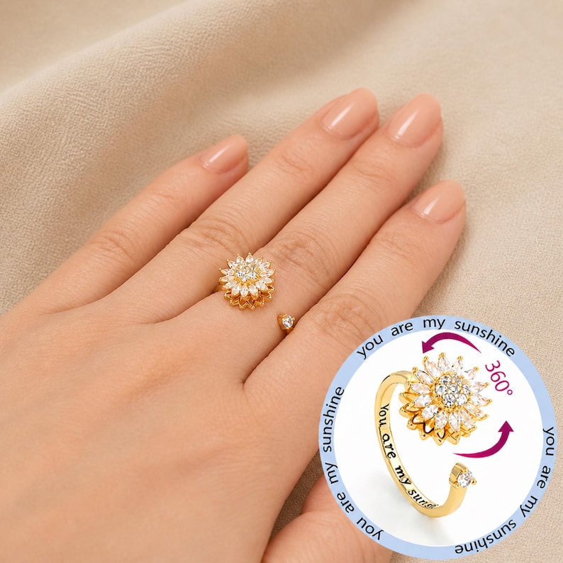 May include: A gold ring with a sunflower design, featuring a central cluster of clear stones and surrounding petals. The ring has a rotating feature and the words "you are my sunshine" are engraved on the band.