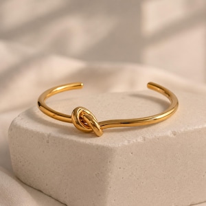 May include: A polished gold-tone cuff bracelet featuring a central knot design. The bracelet has a smooth, reflective surface and an open-ended construction, showcasing a continuous metallic band that loops into a singular knot at the front.