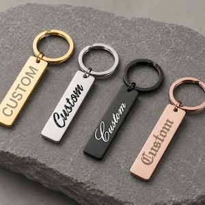 May include: Four rectangular keychains in gold, silver, black, and rose gold. Each keychain has a ring and the word "CUSTOM" engraved on it. The word "CUSTOM" is in a different font on each keychain.