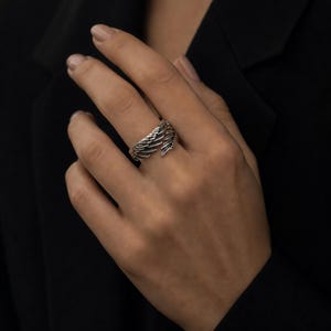May include: A silver-toned ring with a wing design. The ring features detailed feather textures, wrapping around the finger. The ring is worn on a hand, with a dark background.