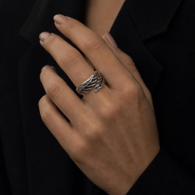 May include: A silver-toned ring with a wing design. The ring features detailed feather textures, wrapping around the finger. The ring is worn on a hand, with a dark background.