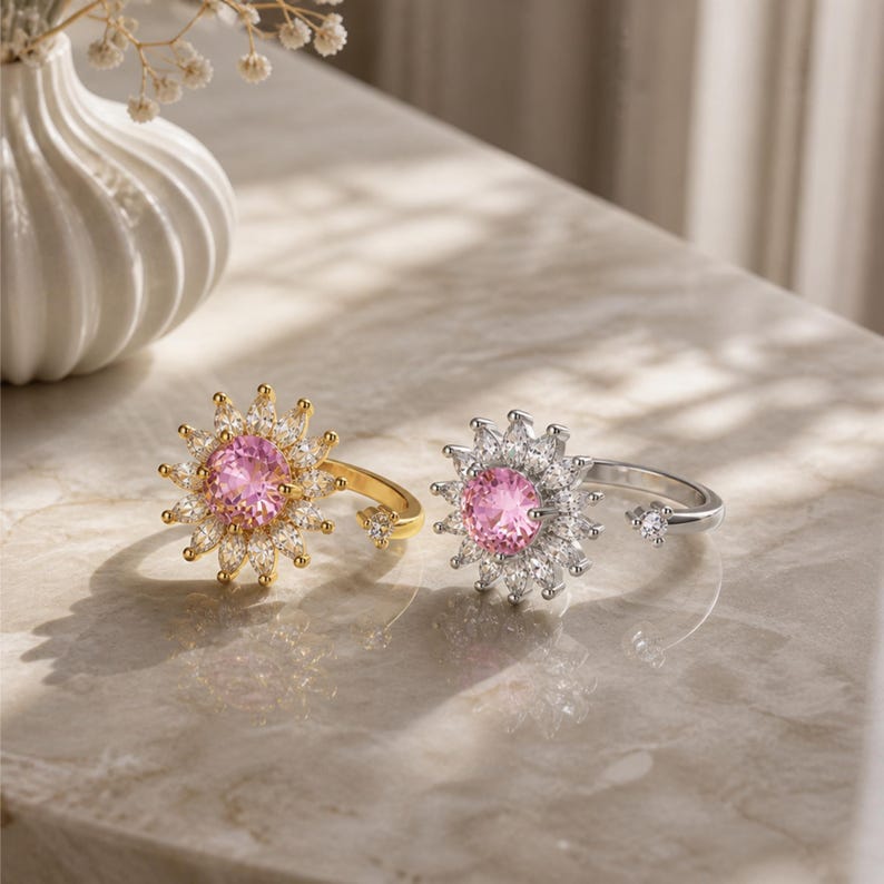 May include: Two rings with a floral design. One is gold-toned with a pink centre stone and white accent stones. The other is silver-toned with a pink centre stone and white accent stones. Both rings are on a marble surface.