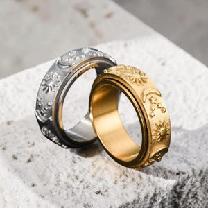 May include: Two rings, one silver and one gold, are shown. The rings have a celestial design with suns, moons, and stars. The gold ring has a shiny finish, while the silver ring has a matte finish. The rings are on a textured, light-coloured surface.