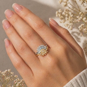 May include: A two-tone ring with a sunburst design. The ring features a gold band and a sun-shaped top with a cluster of small, clear stones and a central yellow stone. The ring is worn on a hand with well-manicured nails.