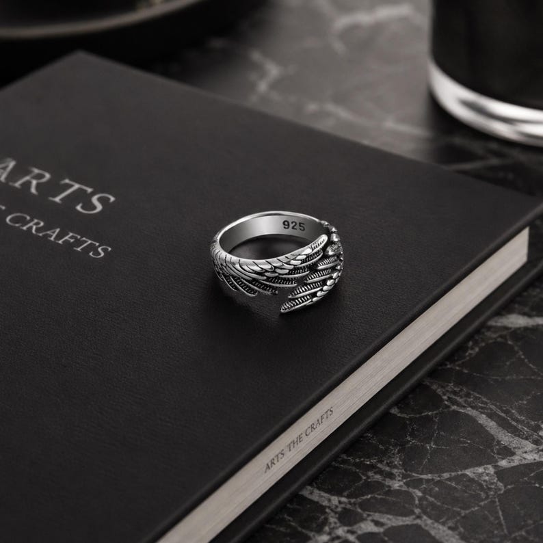 May include: A silver ring with a wing design, marked with "925". The ring is set on a black book with the text "ARTS THE CRAFTS". The ring's wing detail is visible.