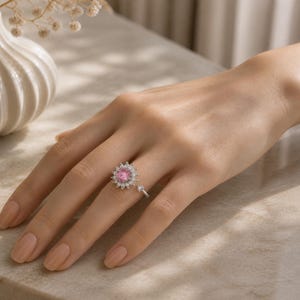 May include: A silver ring with a central pink gemstone, encircled by smaller, clear stones. The ring is displayed on a hand with a neutral manicure, highlighting the jewellery's design. The ring's design is floral-inspired, with a delicate, sparkling appearance.