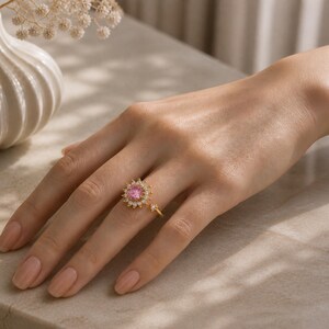 May include: A gold ring with a pink gemstone centre and surrounding white gemstones. The ring is on a hand with painted nails. The ring has a unique design with a small band extending from the main ring.