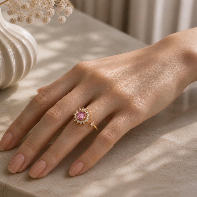 May include: A gold ring with a pink gemstone centre and surrounding white gemstones. The ring is on a hand with painted nails. The ring has a unique design with a small band extending from the main ring.