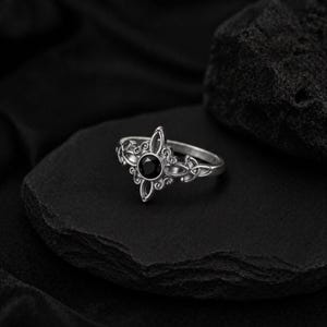 May include: A silver ring with a black gemstone center. The ring features an ornate design with a four-point shape around the gemstone and decorative swirls. The ring is set on a black surface.