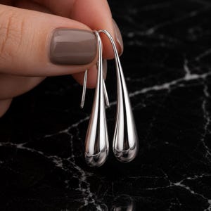 May include: A pair of silver teardrop earrings. The earrings are held against a black and white marble surface. The earrings have a smooth, polished finish and a teardrop shape, with a hook for the ear.