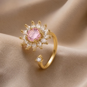 May include: A gold ring with a flower design. The central pink gemstone is surrounded by white gemstones. The open band features a small gemstone detail. The ring is designed to be adjustable.