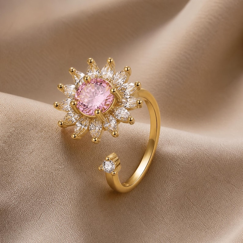 May include: A gold ring with a flower design. The central pink gemstone is surrounded by white gemstones. The open band features a small gemstone detail. The ring is designed to be adjustable.