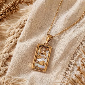 Peut inclure: Un collier pendentif rectangulaire doré arborant un motif de carte de tarot avec l'inscription THE MOON. Le pendentif présente une incrustation irisée blanche et des motifs célestes dorés en relief, incluant lune, étoiles et nuages avec des pierres.