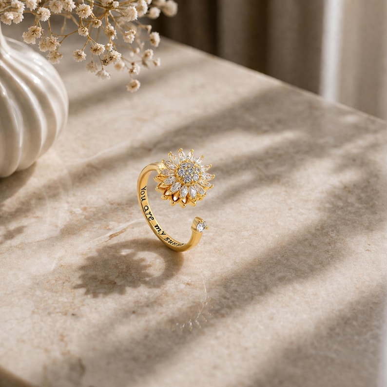 May include: A gold ring featuring a sunflower design, crafted with clear crystals. The band has a small crystal at the end, and the inscription "You are my sunshine" is engraved on the ring.