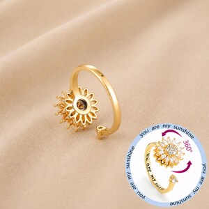 May include: A gold-coloured sunflower ring with a spinning centre. The ring has a delicate design with a band that wraps around the finger. The text "you are my sunshine" is visible on the ring.