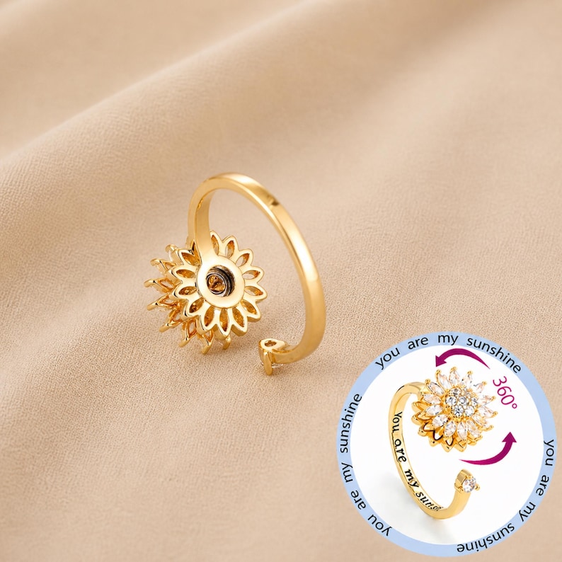 May include: A gold-coloured sunflower ring with a spinning centre. The ring has a delicate design with a band that wraps around the finger. The text "you are my sunshine" is visible on the ring.