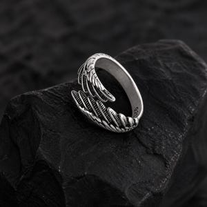 May include: A silver-colored ring with a claw design. The ring features detailed textures and a wrap-around style, with the claw extending from the band. The ring is set against a dark, textured background.