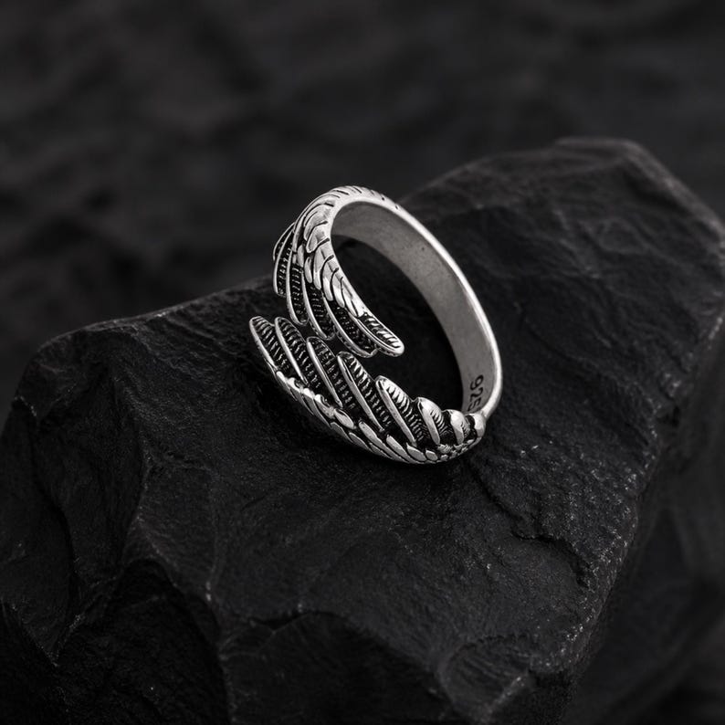 May include: A silver-colored ring with a claw design. The ring features detailed textures and a wrap-around style, with the claw extending from the band. The ring is set against a dark, textured background.