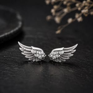 Può includere: Un paio di orecchini a lobo color argento a forma di ali piumate. Ogni ala presenta un design curvo con linee incise dettagliate che rappresentano le piume e un gruppo di piccole pietre rotonde trasparenti incastonate alla base.