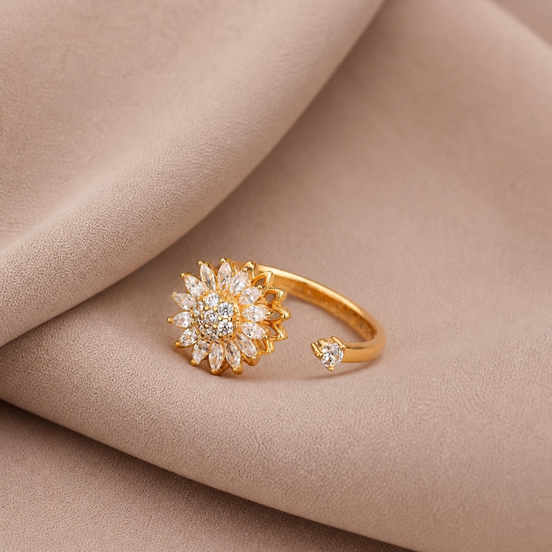 May include: A gold ring with a sunflower design. The ring's centre is decorated with small, round, clear stones, encircled by elongated, clear stones. A small, clear stone accents the open band.