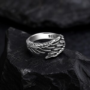 May include: A silver ring with a wing design. The ring is made of polished metal and features intricate feather detailing. The ring is marked with "925" on the inside. The ring is displayed on a dark, textured surface.